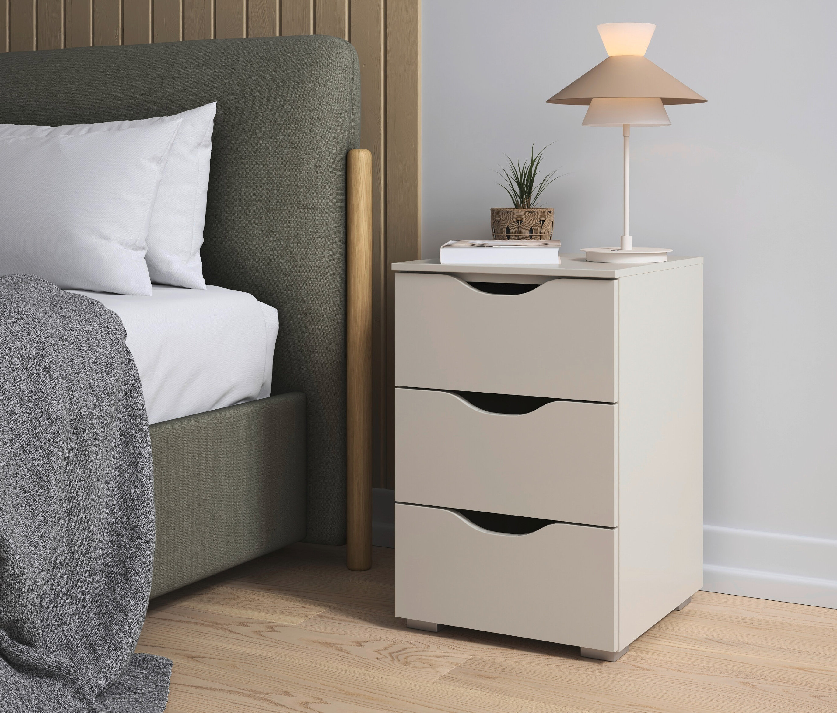 Bett mit grauem Überwurf neben einem rauch Nachttisch »Frieda« mit drei Schubladen in Beige/Champagner, darauf eine Lampe und eine Pflanze.