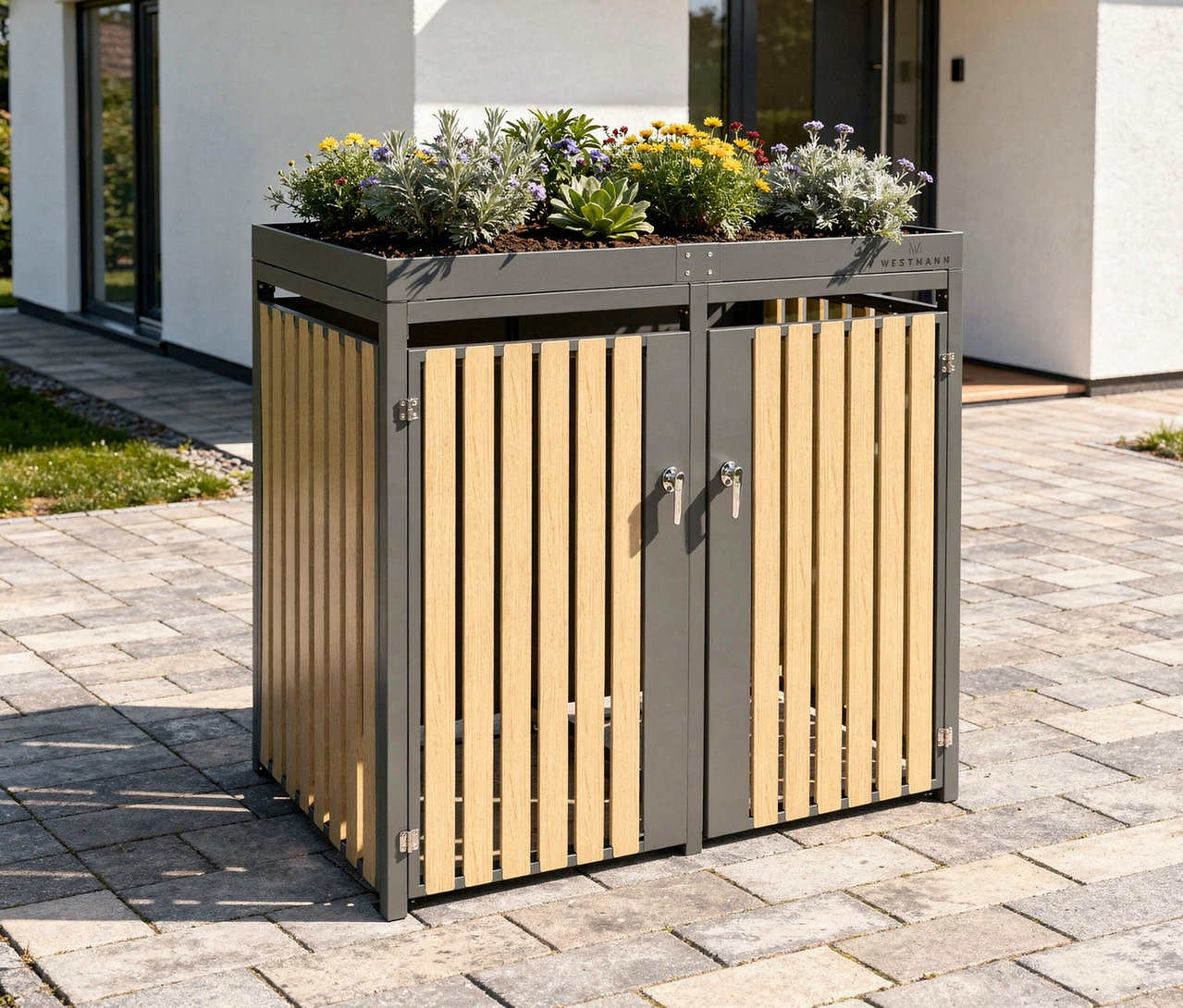 Eine Mülltonnenbox mit Blumenkasten für 2 Tonnen steht auf Pflastersteinen vor einem Haus.
