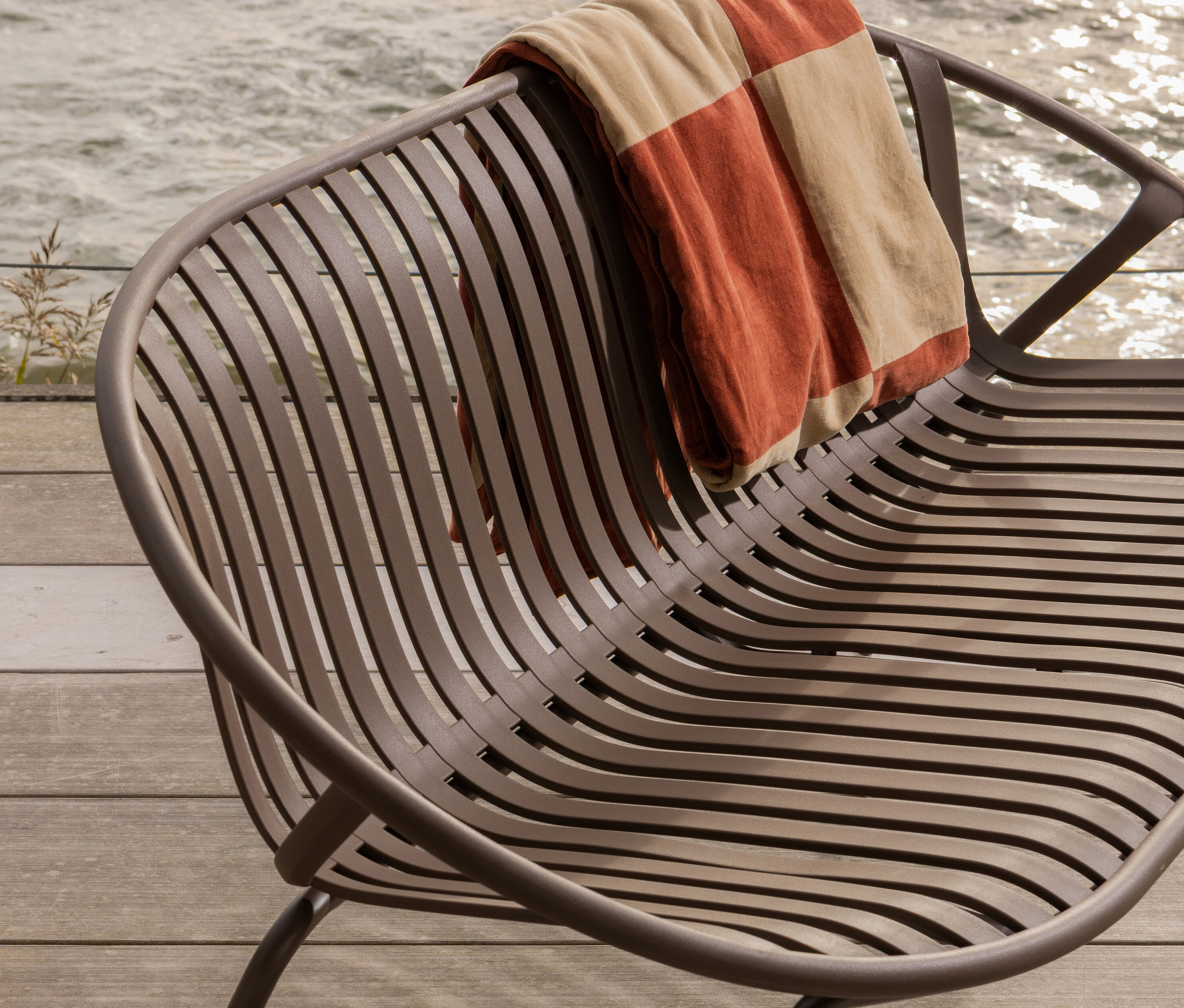 Ein brauner Metallstuhl mit einer gestreiften Decke steht auf einer Holzterrasse am Wasser.