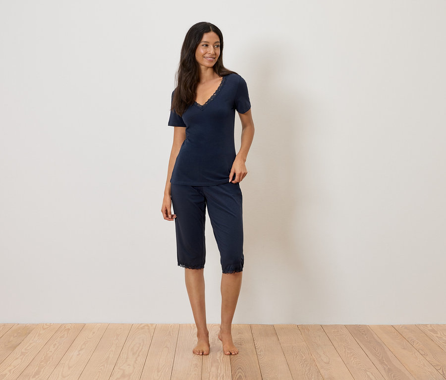 Frau steht barfuß auf einem Holzboden und trägt einen marineblauen Pyjama mit Spitzenbesatz.