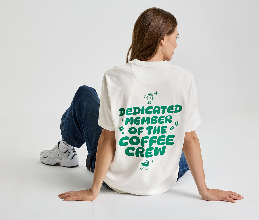 Frau sitzt auf dem Boden, trägt ein T-Shirt mit der Aufschrift "Dedicated Member of the Coffee Crew", dazu Jeans und Turnschuhe.