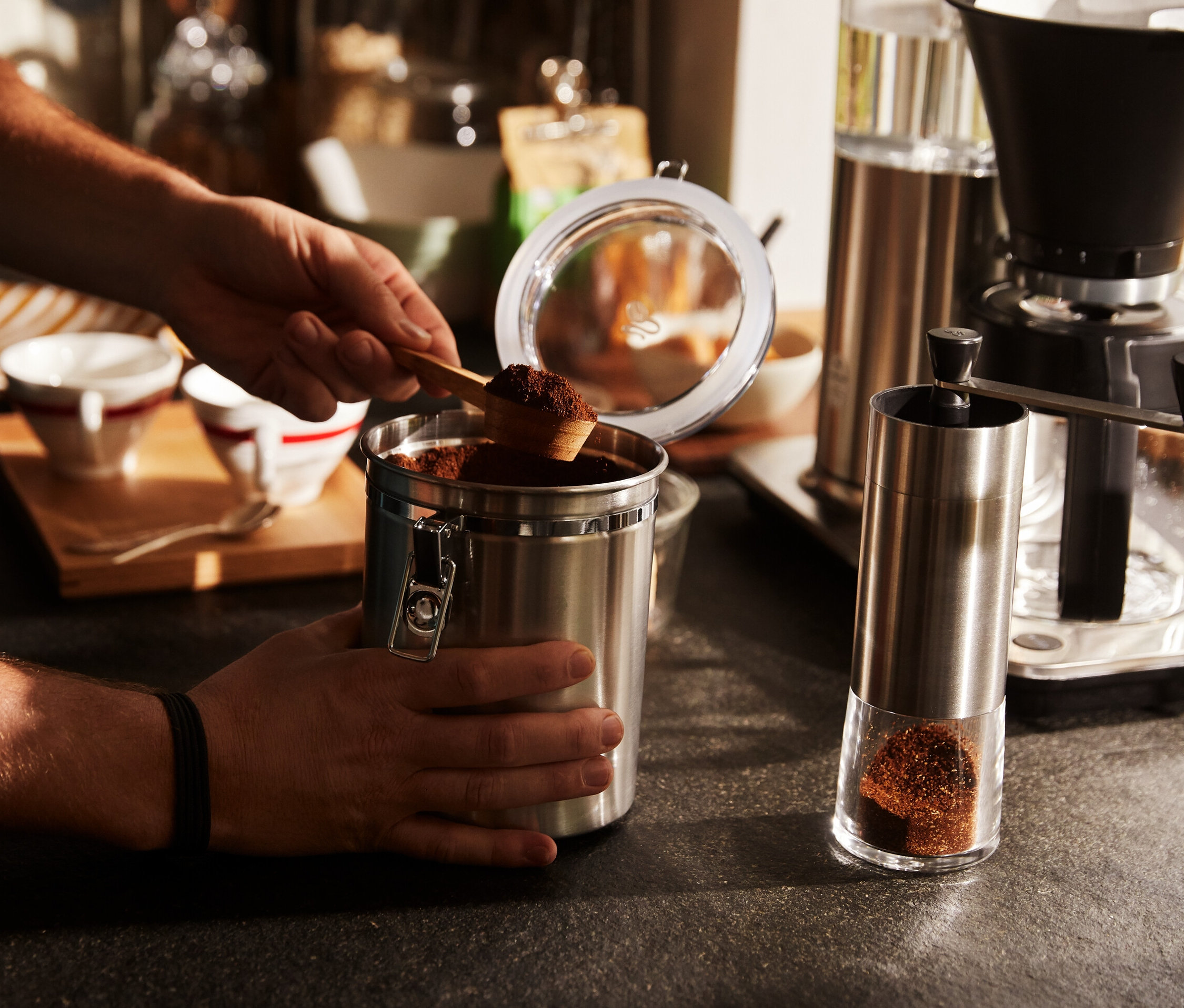 Mann schöpft mit Löffel Kaffee aus Blechdose. Daneben steht eine silberne, manuelle Kaffeemühle.