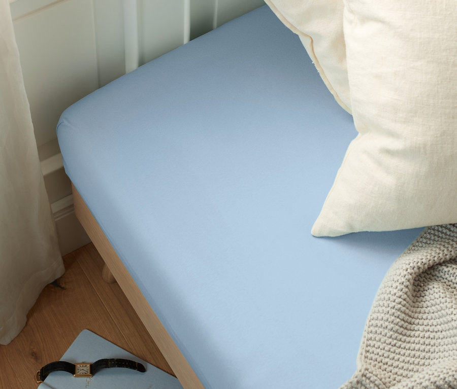 Blaues Jersey-Spannbettlaken in Normalgröße auf einem Bett mit weißen Kissen und einer grauen Decke.