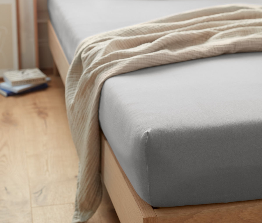 Graues Jersey-Spannbettlaken auf einem Bett mit Holzrahmen und weißen Kissen.