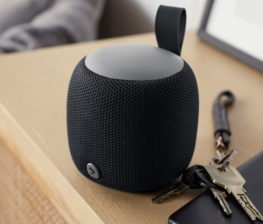 Ein kleiner, schwarzer Fabric-Bluetooth®-Lautsprecher steht auf einem Holztisch neben einem Mobiltelefon und Schlüsseln.