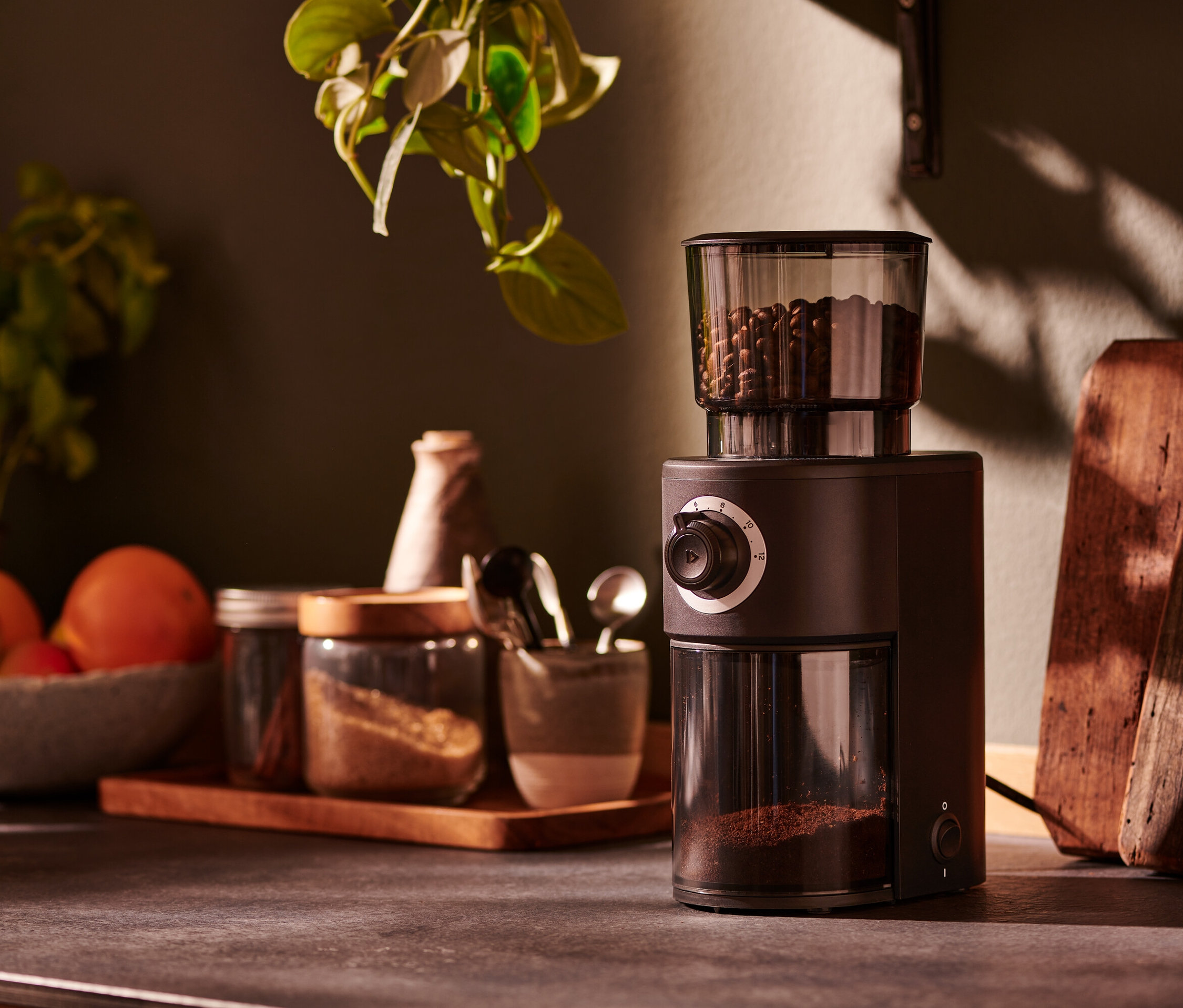 Eine braune, elektrische Kaffeemühle steht auf einer Küchentheke neben Gläsern mit Gewürzen.