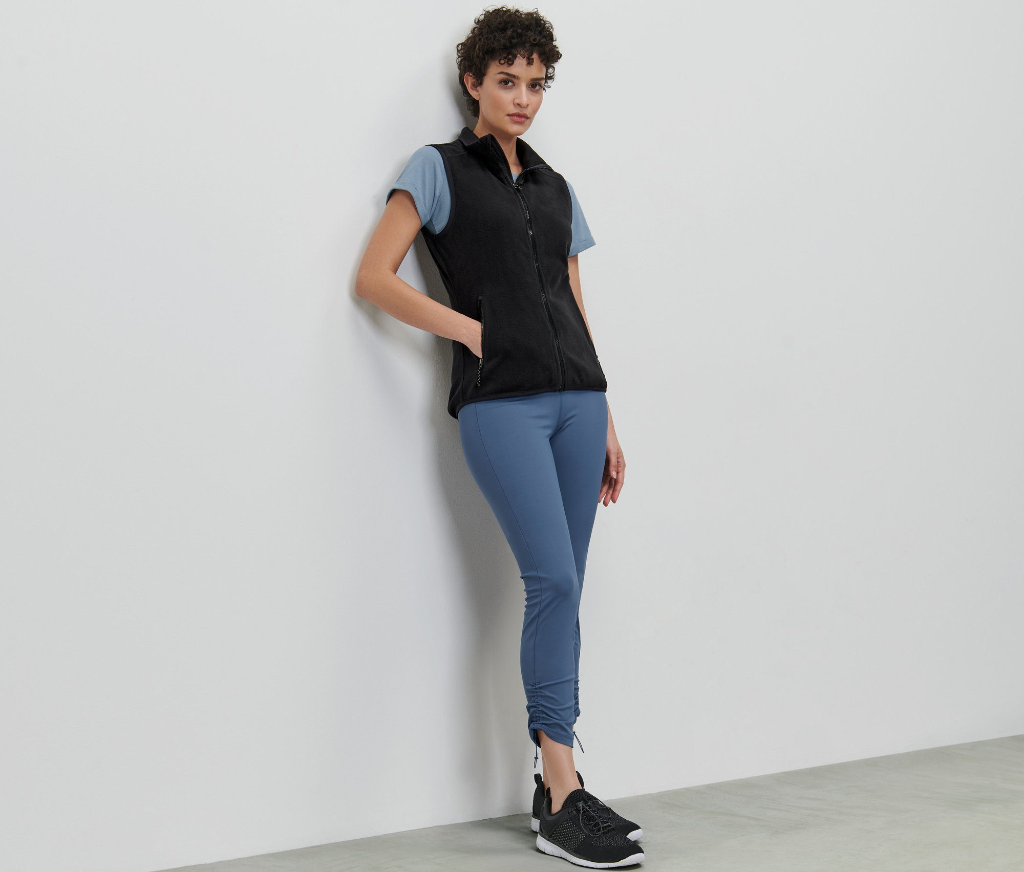 Eine Frau lehnt an einer Wand und trägt eine Fleece-Weste, ein T-Shirt und Leggings.