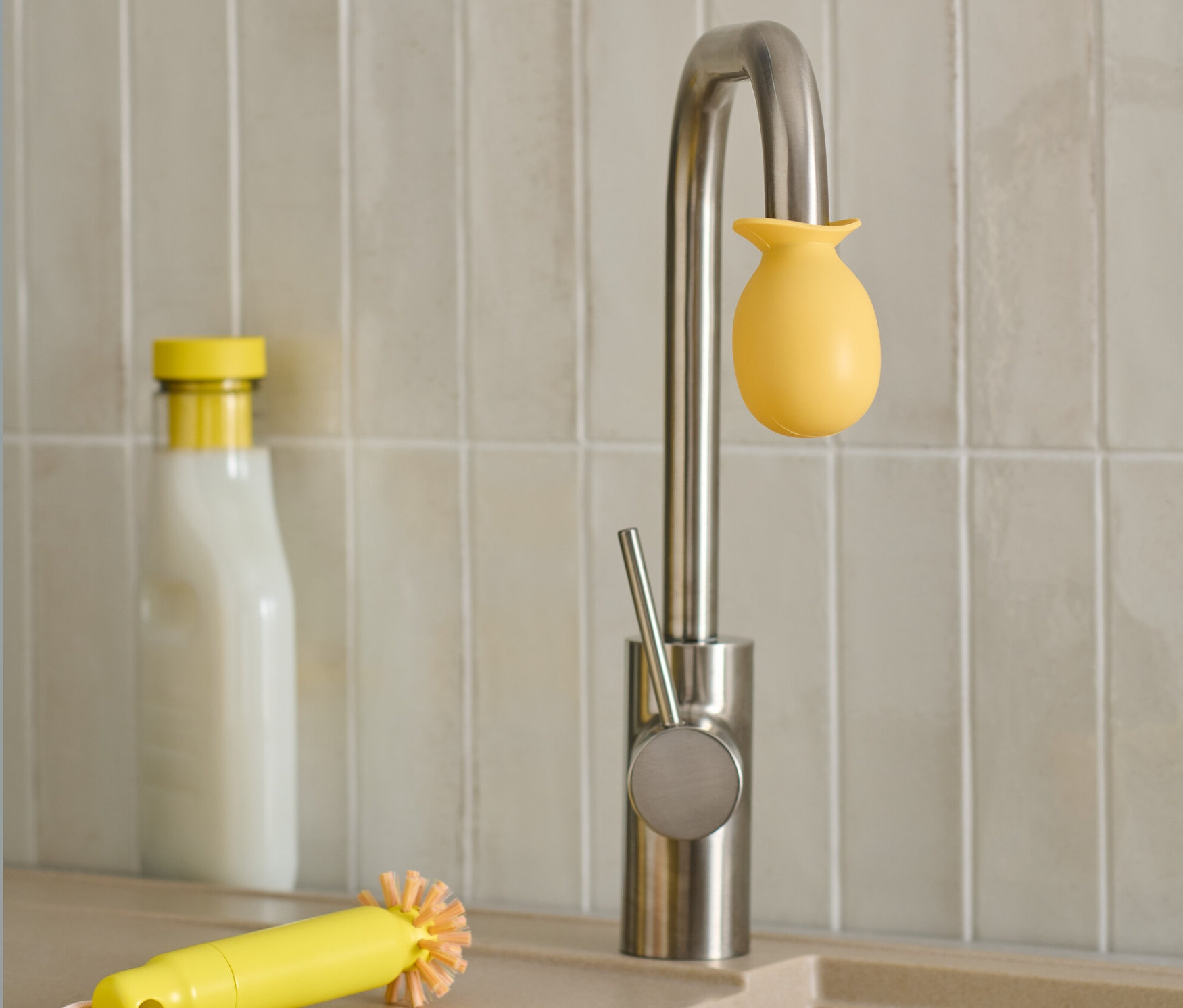 Auf einer Edelstahl-Armatur befindet sich ein gelber Wasserhahn Entkalker-Aufsatz. Im Hintergrund stehen eine Flasche und eine gelbe Bürste.