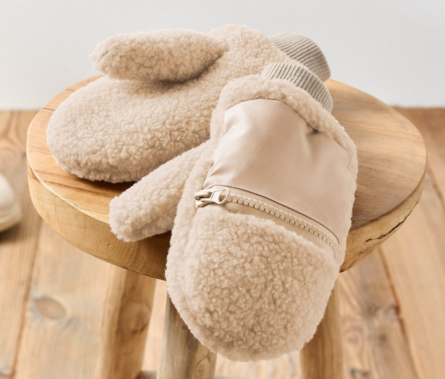 Zwei Teddy-Fäustlinge liegen auf einem Holzhocker. Im Hintergrund sind beige Stiefel zu sehen.