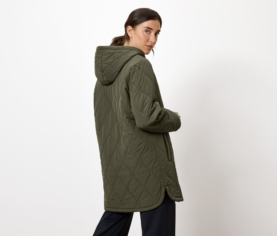 Eine Frau blickt über ihre Schulter und trägt einen olivgrünen Steppmantel mit Kapuze.