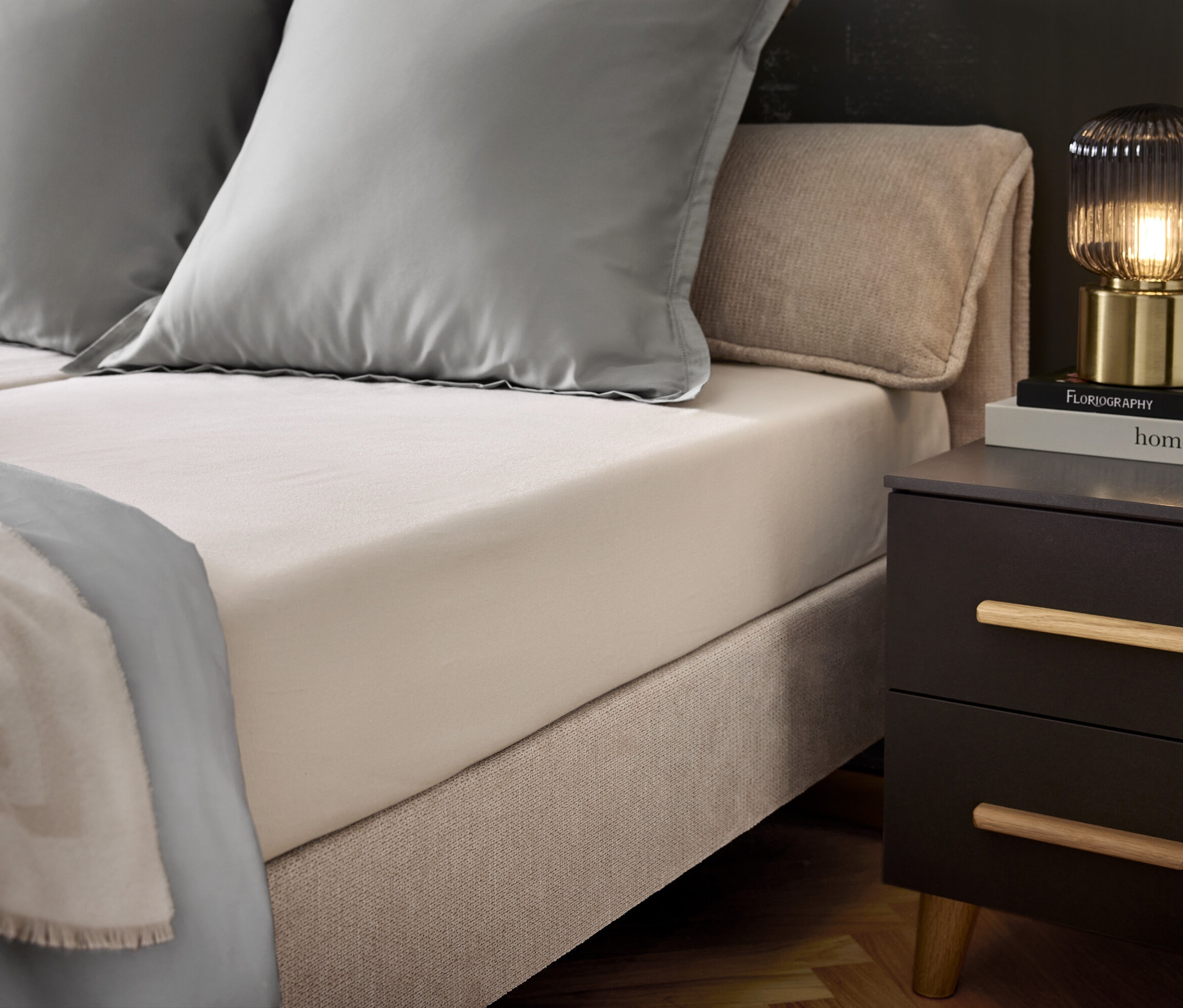 Schlafzimmer mit Bett, zwei grauen Kissen und einer grauen Tagesdecke. Auf dem Nachttisch stehen eine Lampe und Bücher.