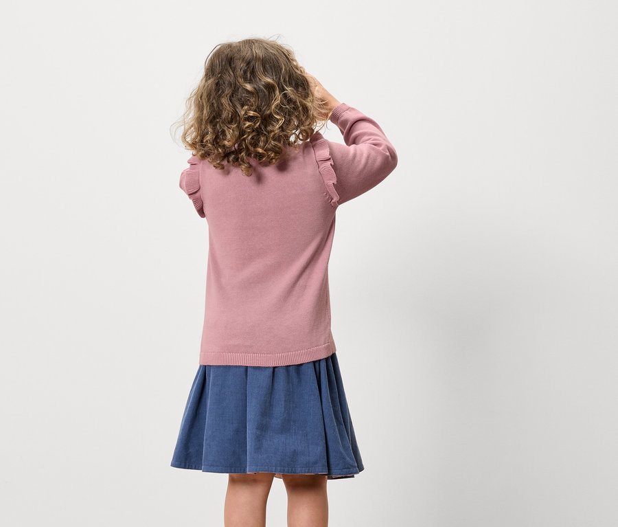 Ein Mädchen mit lockigem Haar steht mit dem Rücken zur Kamera und trägt einen rosa Pullover und einen blauen Rock.