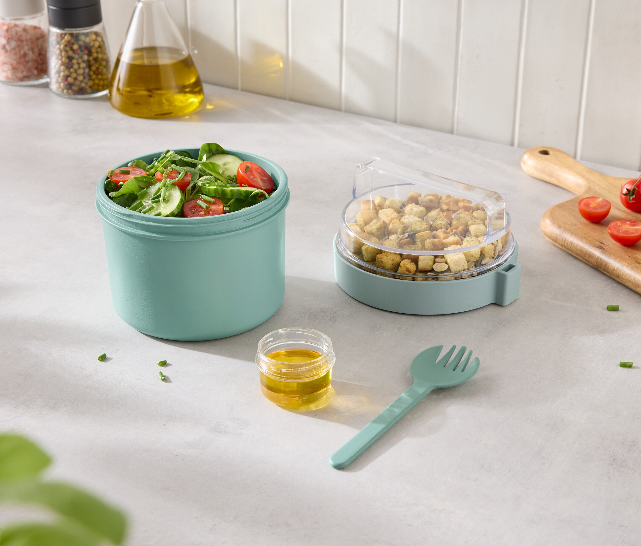 Gemischter Salat in türkisblauem Behälter mit Croutons in einem Behälter mit transparentem Deckel und Dressing und Gabel daneben.