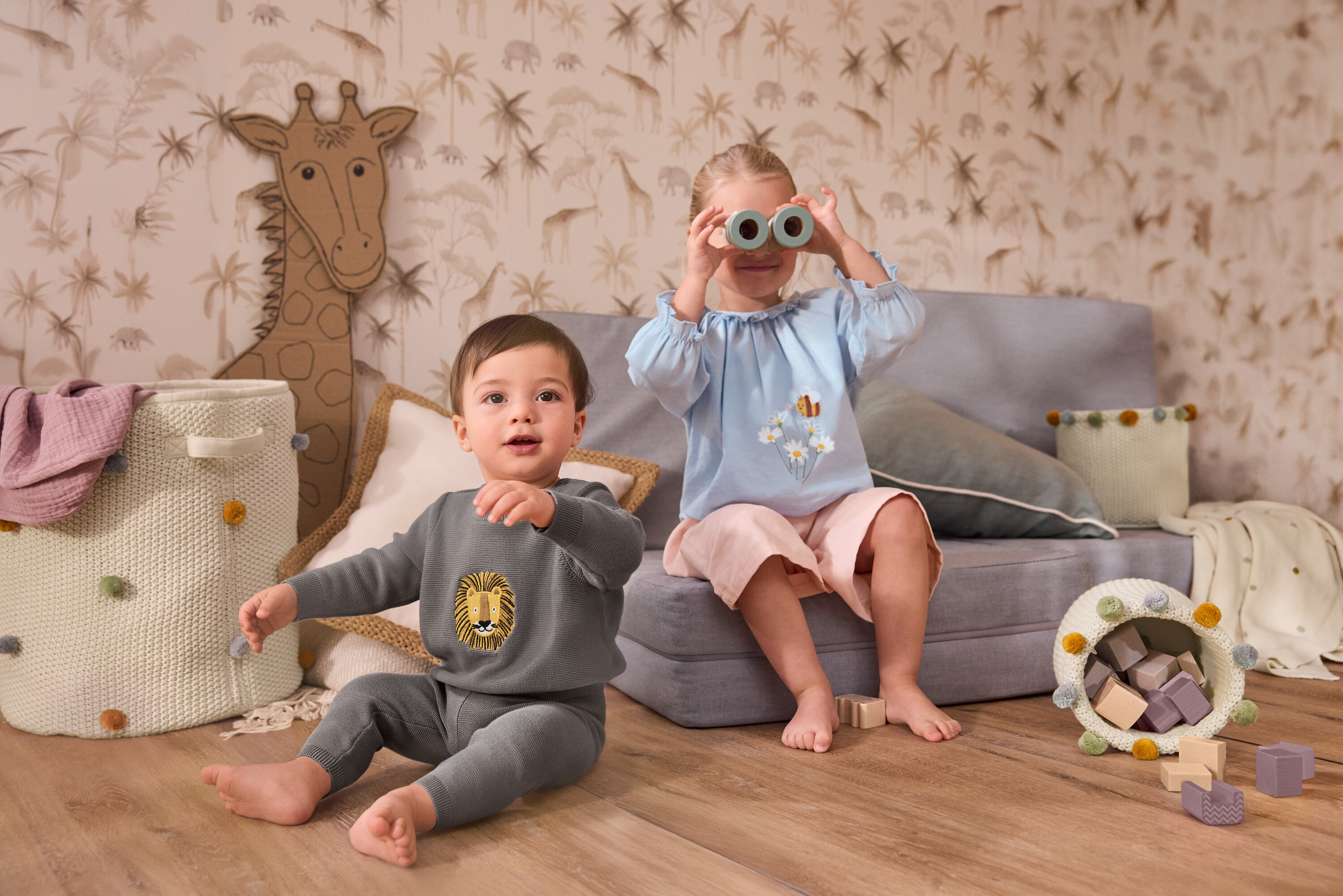 Ein Kleinkind sitzt neben einem großen Kinderzimmer-Aufbewahrungskorb. Ein Mädchen sitzt auf einem grauen Sofa und hält eine Strick-Kuscheldecke für Babys vor ihr Gesicht. Sie trägt eine Kinder-Chambray-Bluse und eine Kinder-Cord-Culotte.
