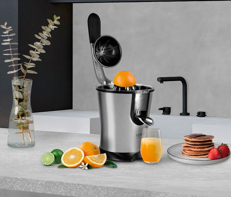 Eine Zitruspresse CP200 mit Orange darauf, neben einem Glas Orangensaft und Pfannkuchen.
