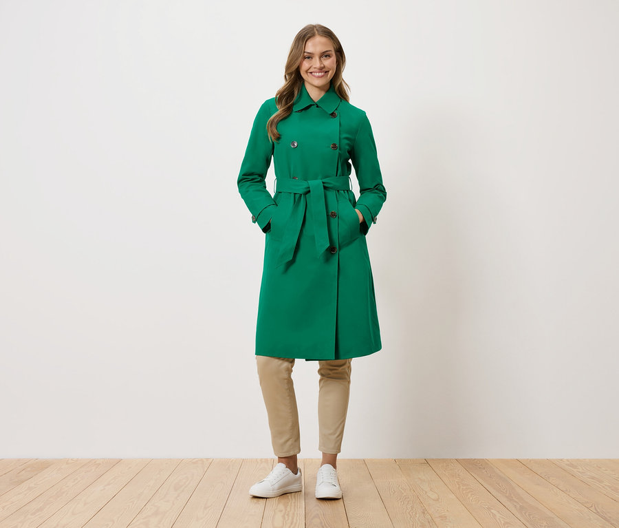Eine Frau steht mit ihren Händen in den Taschen eines grünen Trenchcoats.
