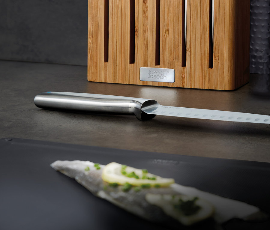 Ein Messer mit Edelstahlgriff und gelochter Klinge liegt auf einem schwarzen Schneidebrett, daneben ein Messerblock aus Holz.