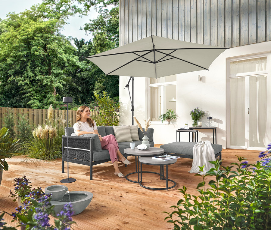 Eine Frau sitzt auf einem komfortablen Gartensofa mit separater Fußlehne. Eine graue Solar-Akku-Stehleuchte steht neben ihr. Im Vordergrund befindet sich ein Solar-Gartenbrunnen.