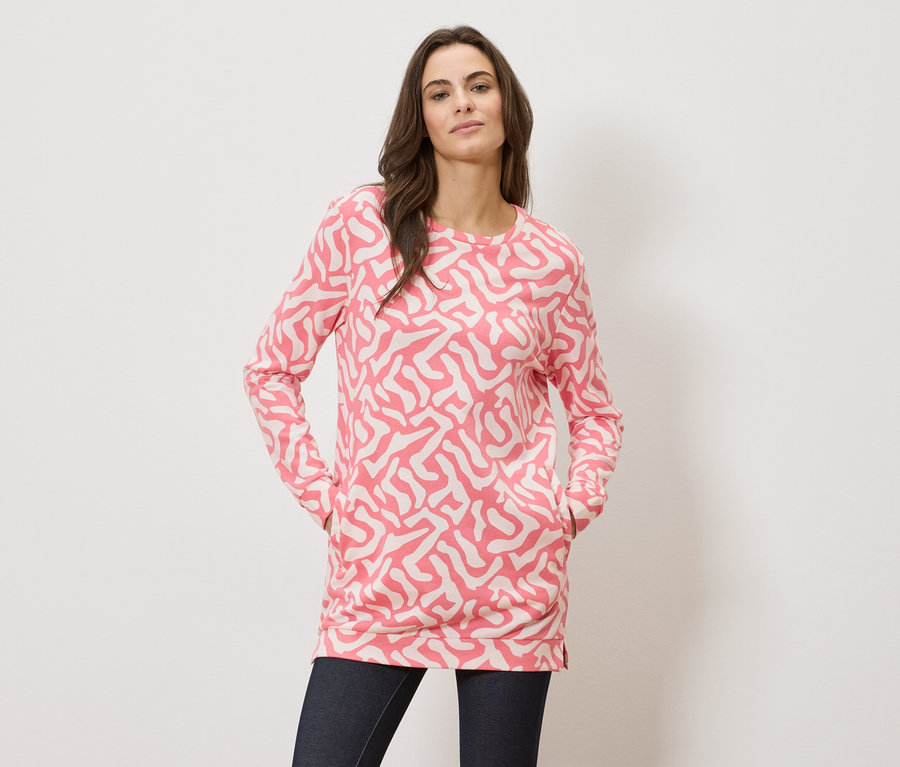 Frau mit ihren Händen in den Taschen, sie trägt ein langärmliges Oberteil mit pinkfarbenem Muster und dunkle Jeans.