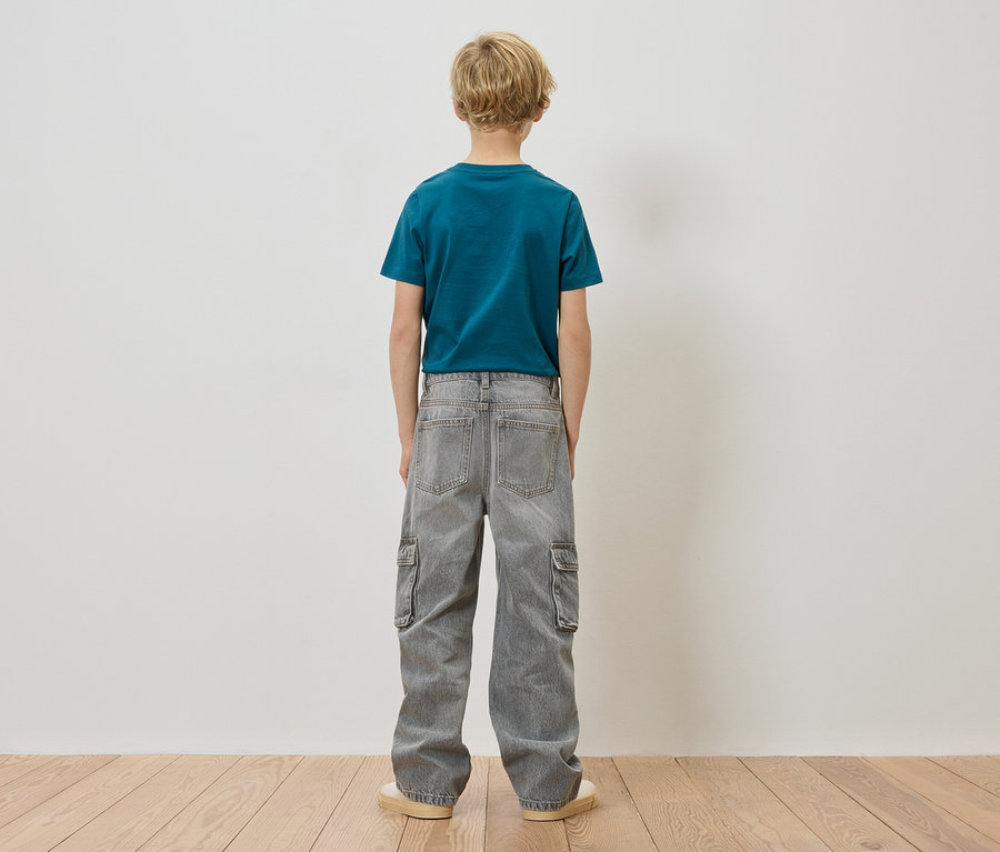 Ein Kind mit blonden Haaren steht mit dem Rücken zur Kamera, es trägt ein blaugrünes T-Shirt und graue Cargo-Jeans.