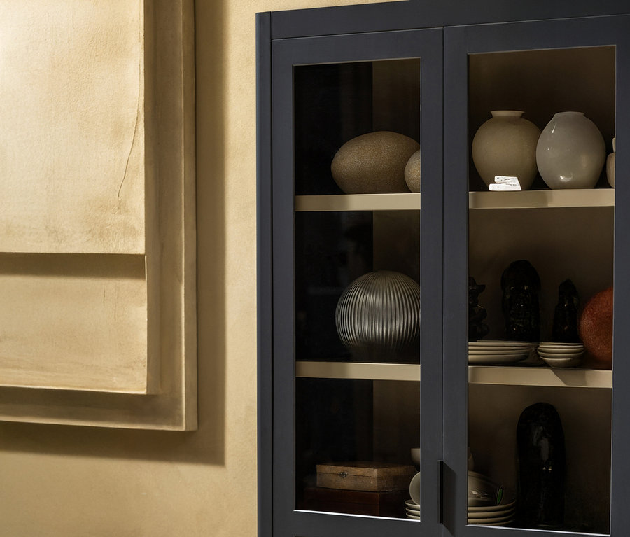 Nahaufnahme eines geschlossenen Glasschranks mit dekorativen Vasen, Tellern und Skulpturen auf mehreren Regalen, neben einem beigen Wandkunstwerk.