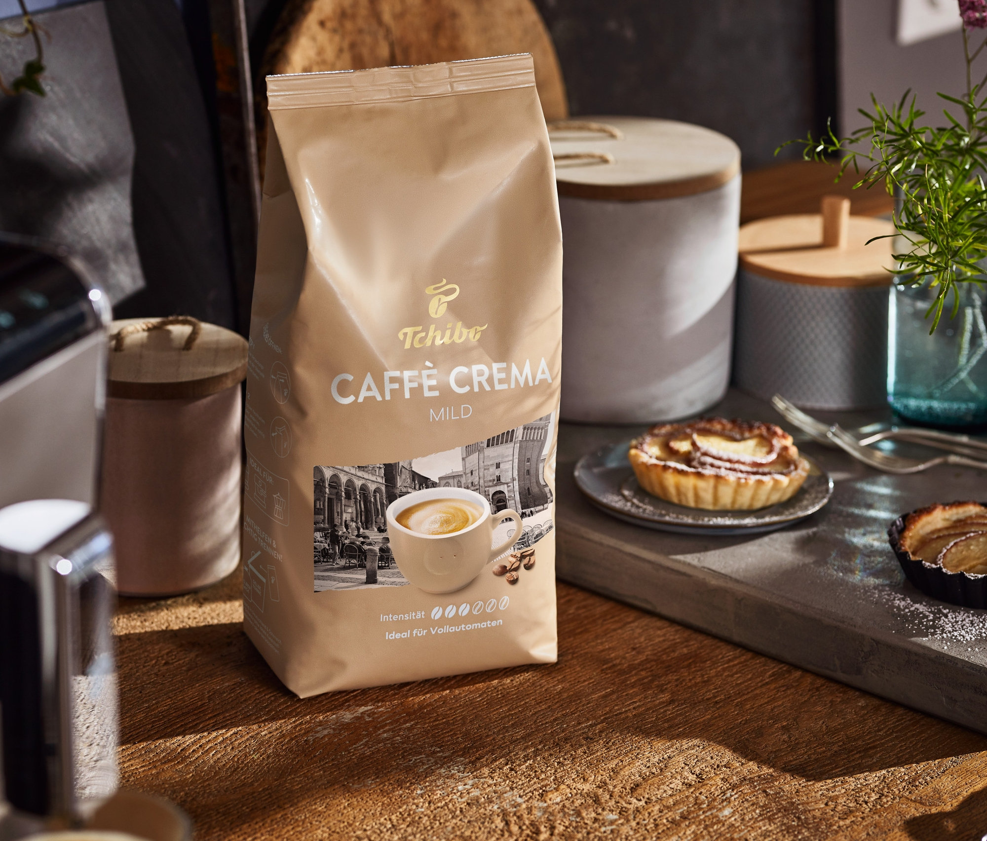 Auf einem Holztisch stehen eine Packung Caffè Crema Mild, eine Kaffeemaschine und Teller mit Kuchen.