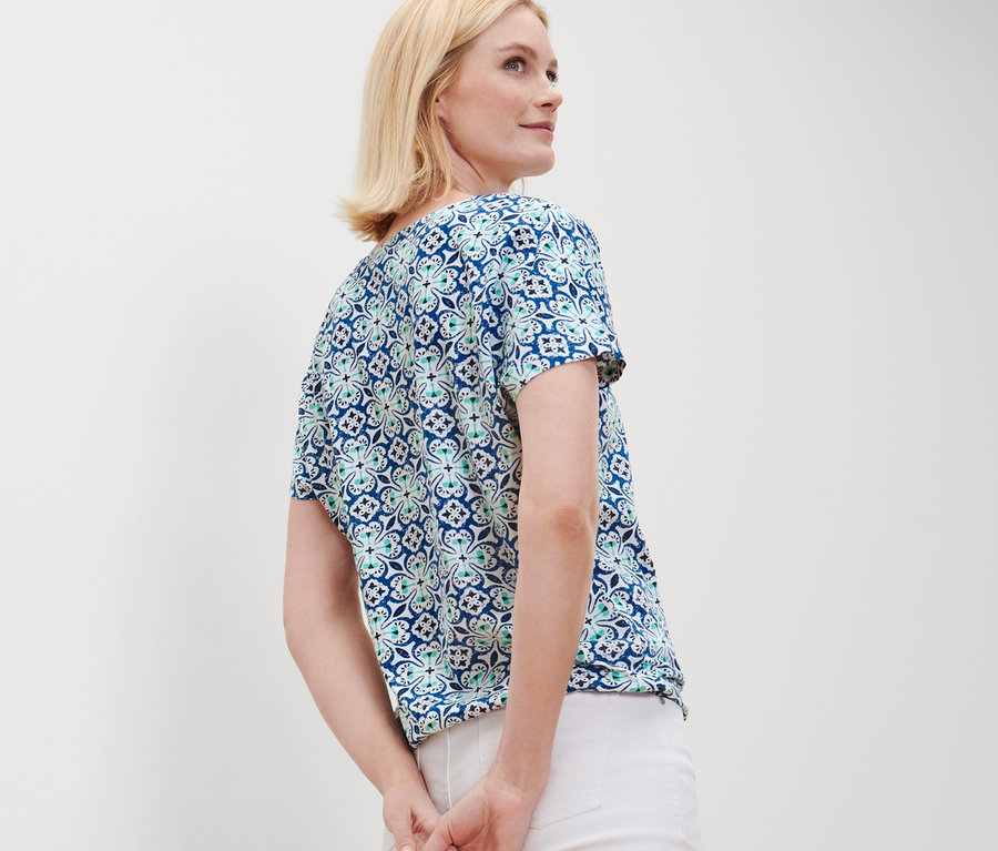 Blonde Frau posiert in einem Blusenshirt mit Alloverprint und weißen Hosen.