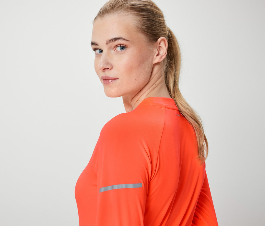 Blonde Frau schaut über die Schulter und trägt ein orangefarbenes Langarmshirt.