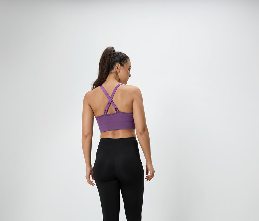 Eine Frau mit braunen, zu einem Pferdeschwanz zusammengebundenen Haaren steht mit dem Rücken zur Kamera und trägt einen lila Sport-BH und schwarze Leggings.