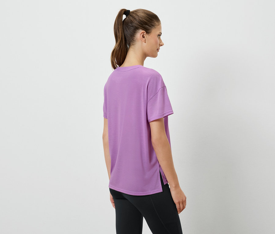 Frau in Lavendel T-Shirt und schwarzen Leggings steht mit dem Rücken zur Kamera.