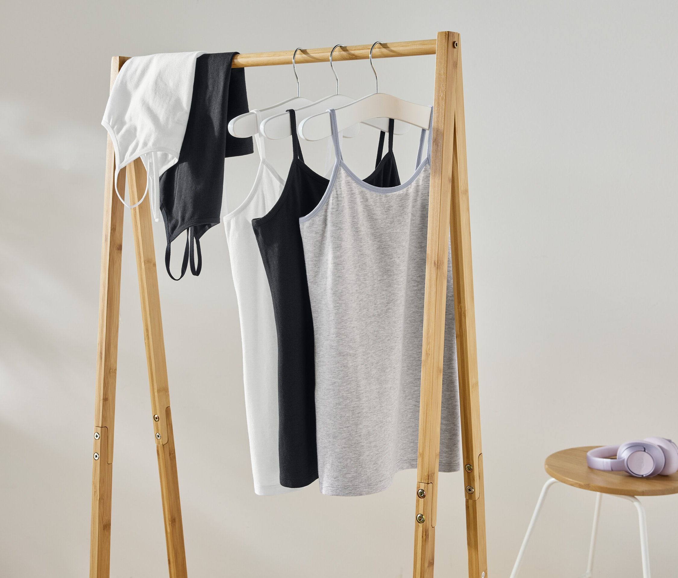 Auf einem hölzernen Kleiderständer hängen ein weißes Top, ein schwarzes Top, ein weißes Trägertop, ein schwarzes Trägertop und ein graues Trägertop. Auf einem Hocker daneben liegen lilafarbene Kopfhörer.