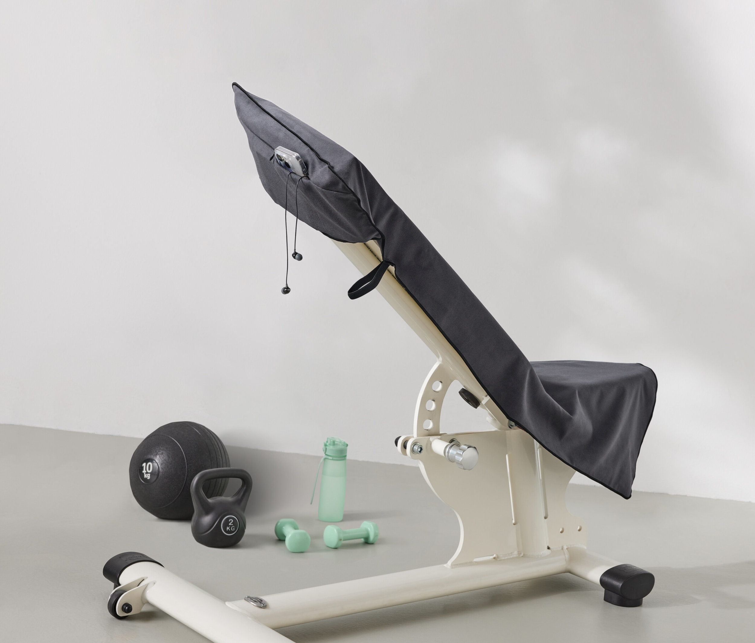 Eine Hantelbank ist mit einem grauen Tuch bedeckt. Neben der Bank liegen ein Medizinball, eine Kettlebell, eine Wasserflasche und Hanteln.