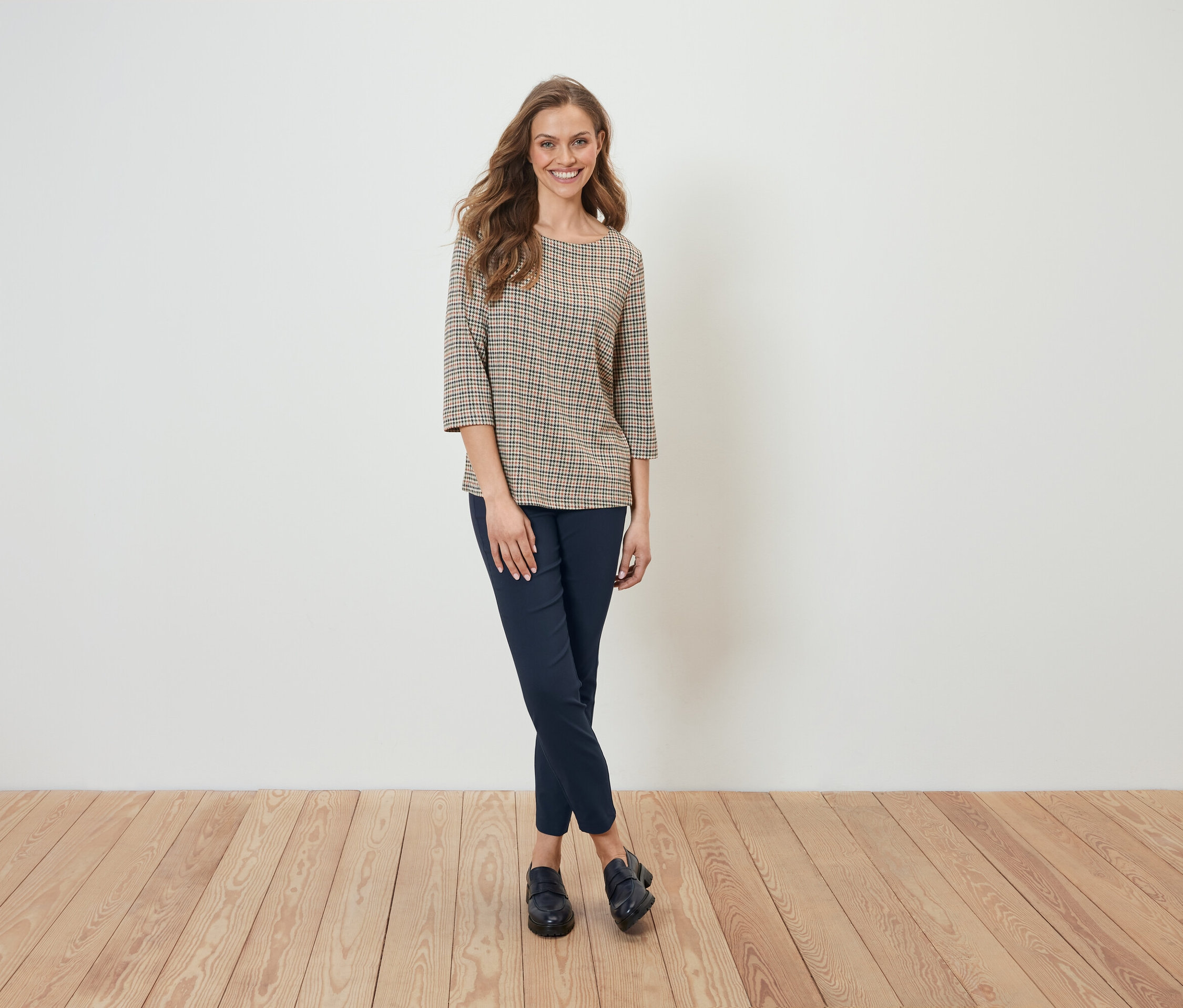 Eine Frau in einer karierten Bluse und dunkelblauen Hosen steht vor einer weißen Wand.