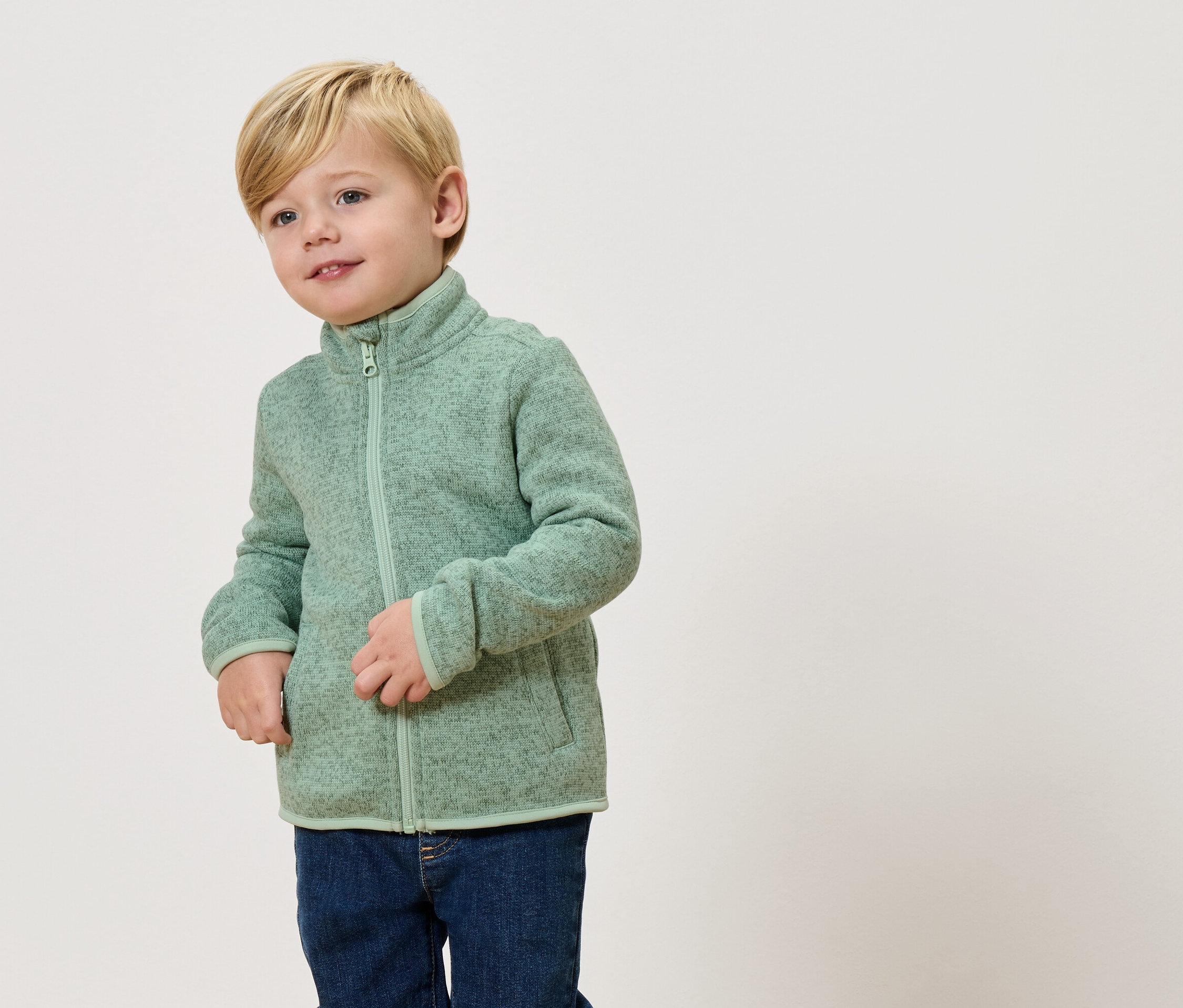 Ein blonder Junge trägt eine hellgrüne Fleecejacke und Jeans.