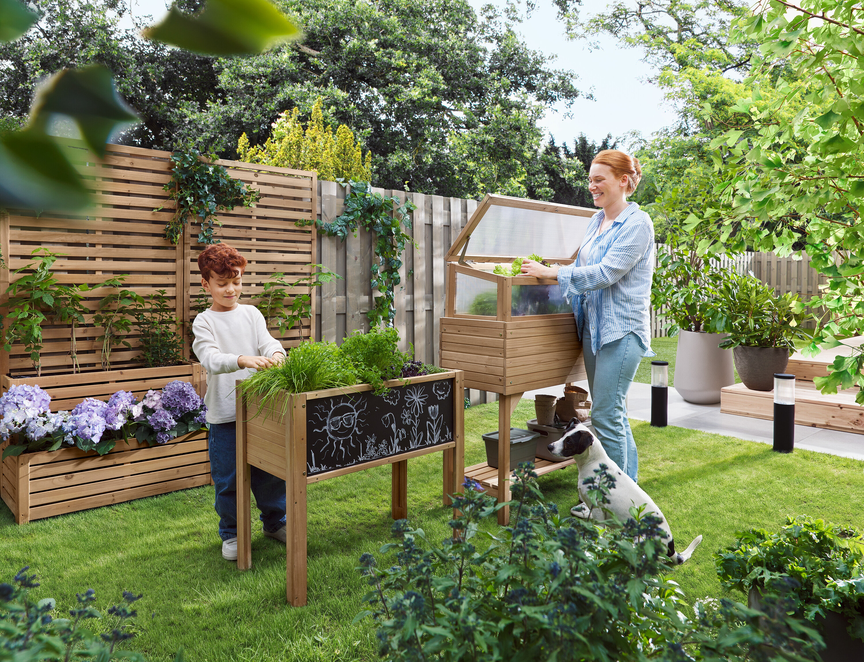 Eine Frau öffnet ein Frühbeet, während ein Kind neben einem Hochbeet steht. Ein Hund sitzt im Gras.