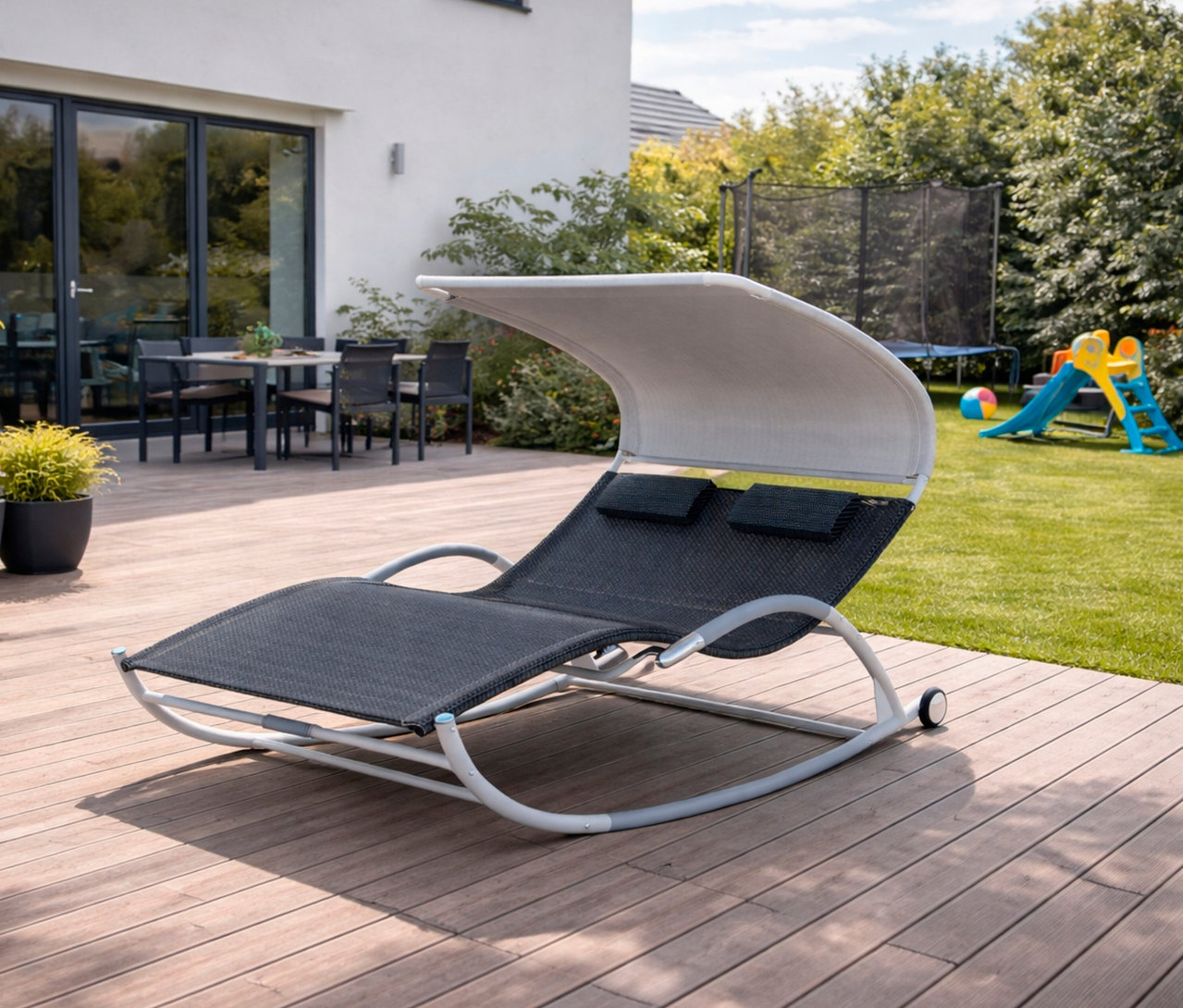Eine Leco Doppelschaukelliege mit Rädern steht auf einer Holzterrasse. Im Hintergrund sind ein Haus, ein Trampolin und eine Rutsche zu sehen.