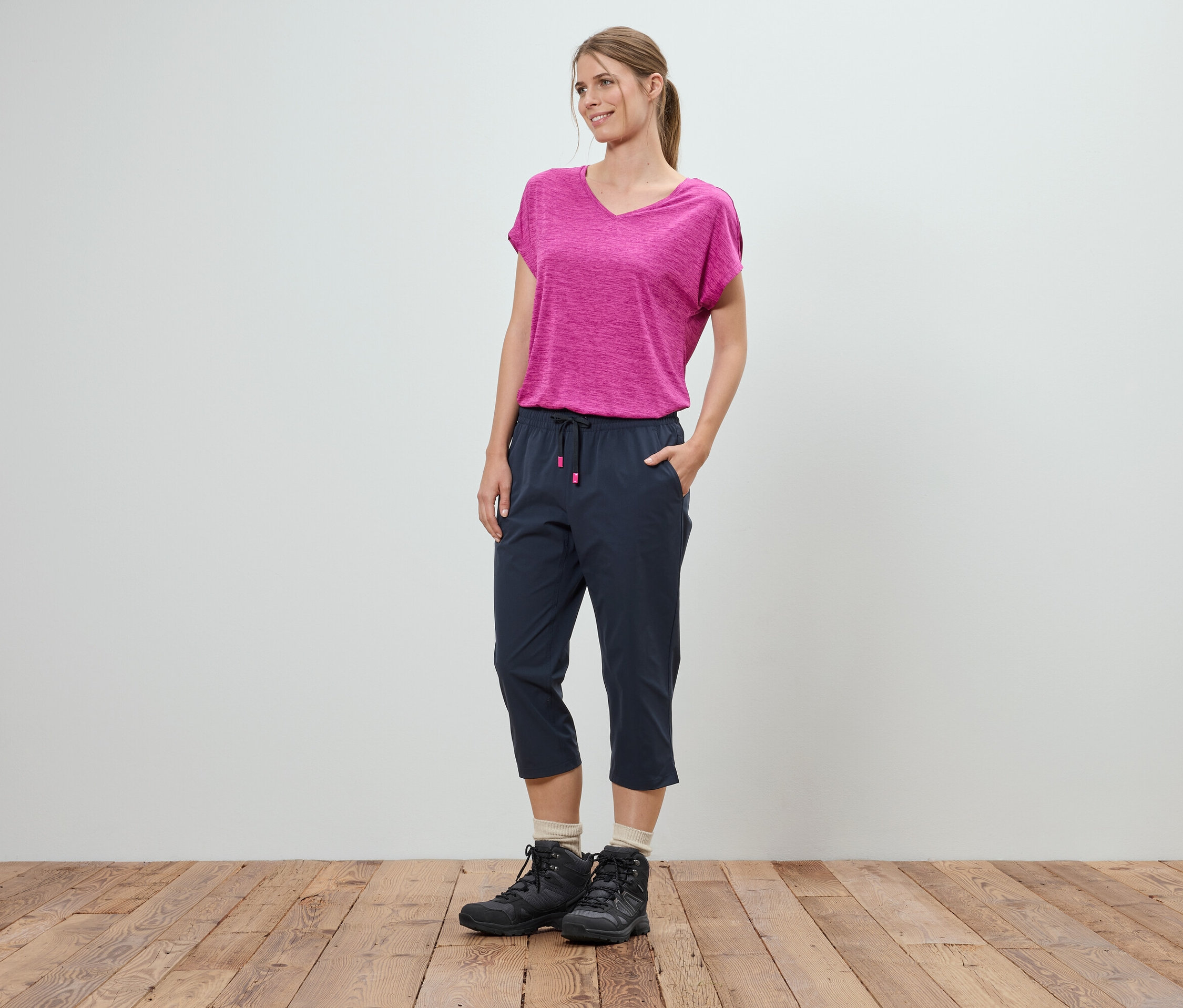 Eine Frau steht mit den Händen in den Taschen vor einer weißen Wand. Sie trägt ein pinkfarbenes T-Shirt, blaue Dreiviertelhosen und schwarze Wanderschuhe.