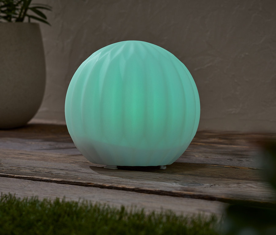 Eine leuchtend türkisfarbene kugelförmige Lampe steht auf einem Holztisch, neben einem Topf mit einer Pflanze.