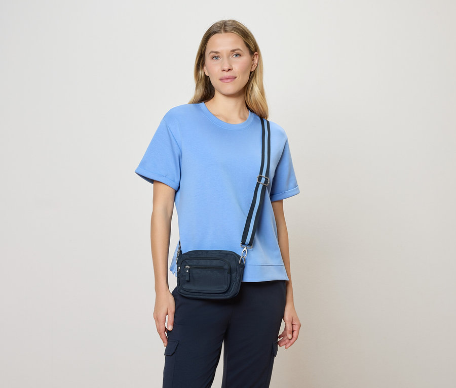 Frau mit hellbraunem Haar, hellblauem T-Shirt und marineblauer Umhängetasche mit marineblauem Gurt vor einem cremefarbenen Hintergrund.