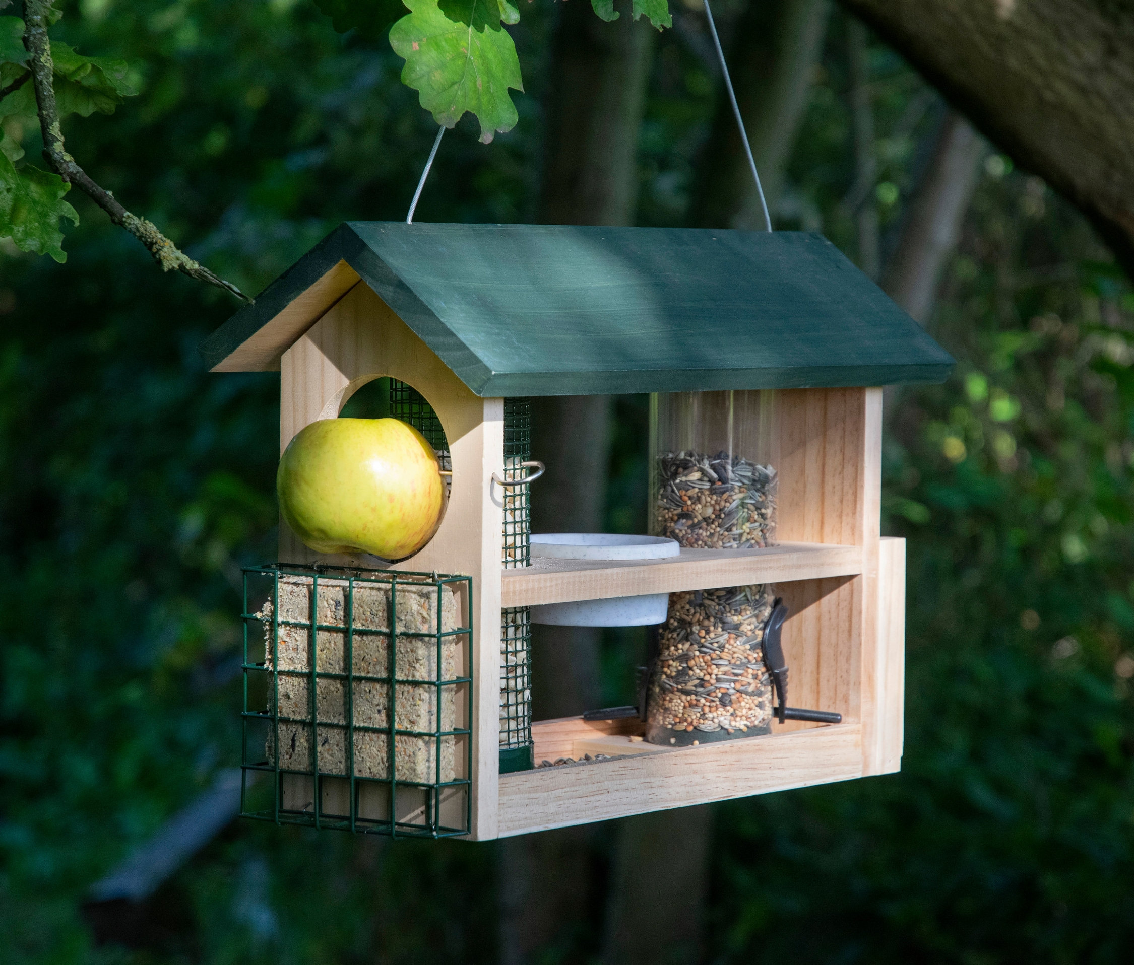 Ein hölzernes 7-in-1-Futterhaus hängt an einem Baum und ist mit Äpfeln, Talg und Vogelfutter gefüllt.