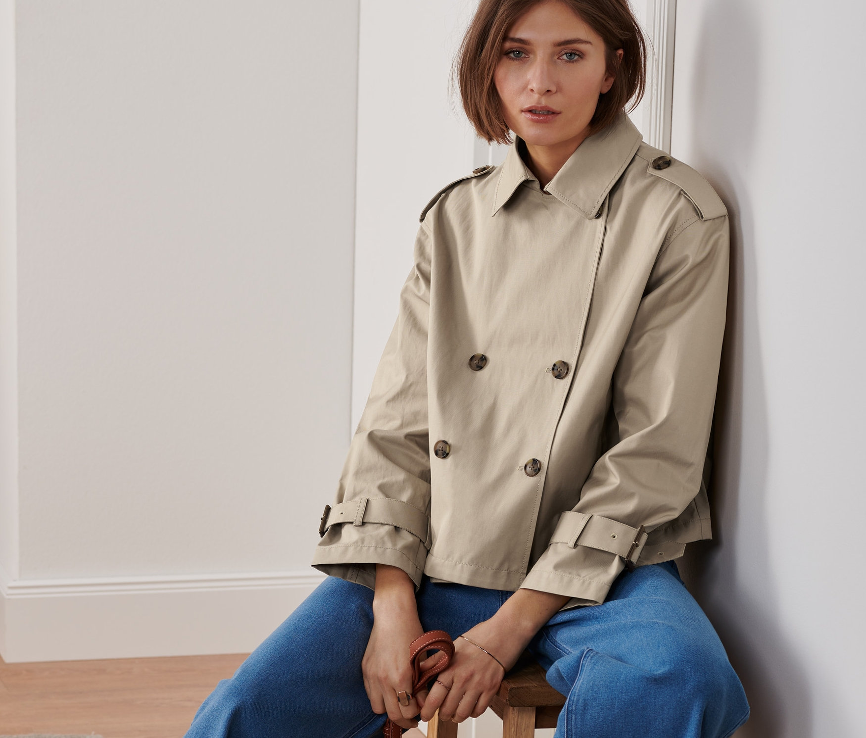 Frau sitzt auf einem Hocker und trägt einen kurzen Trenchcoat und hält eine braune Handtasche.