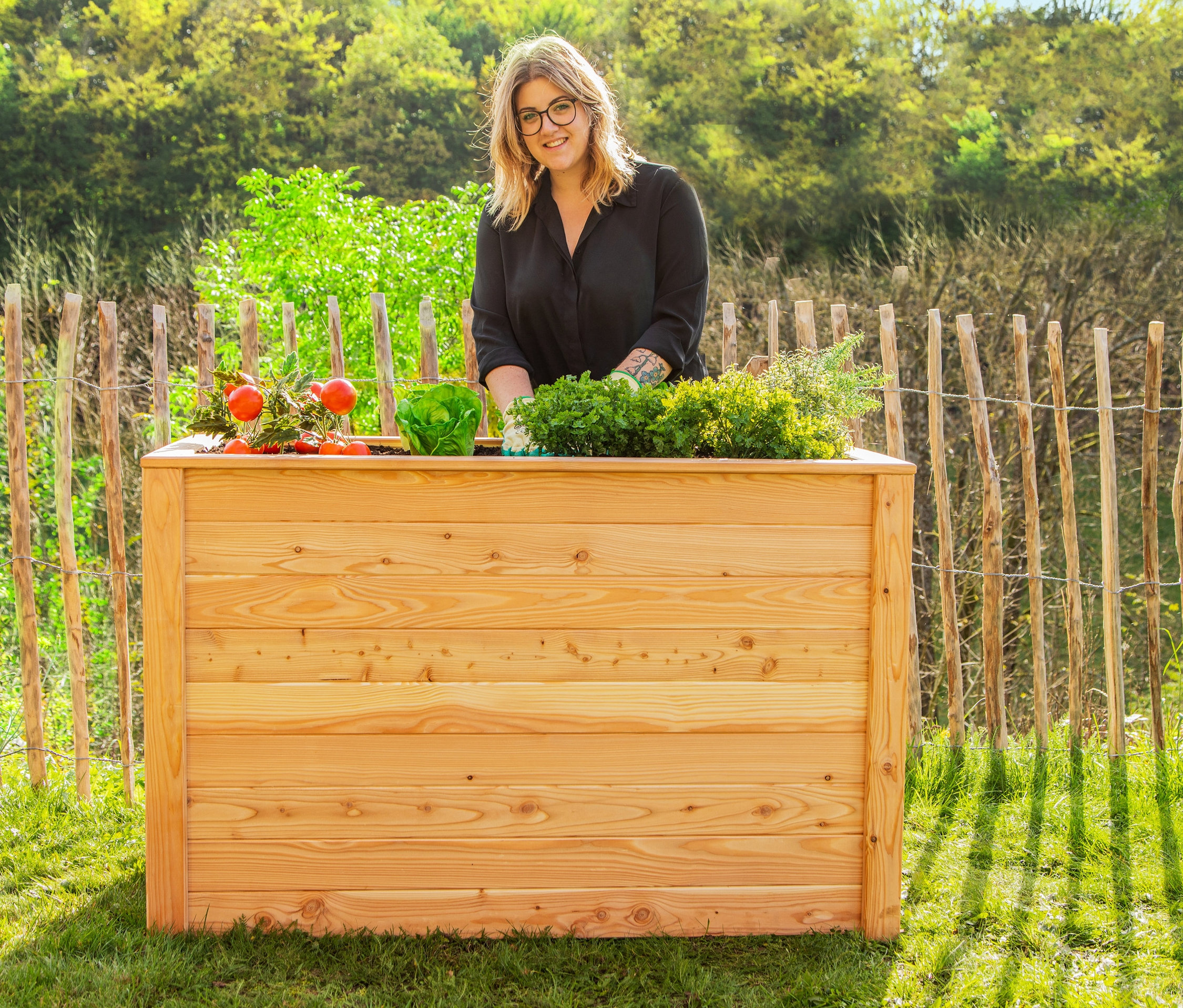 Eine Frau steht hinter einem 130 cm langen Hochbeet aus Lärchenholz und gärtnert.