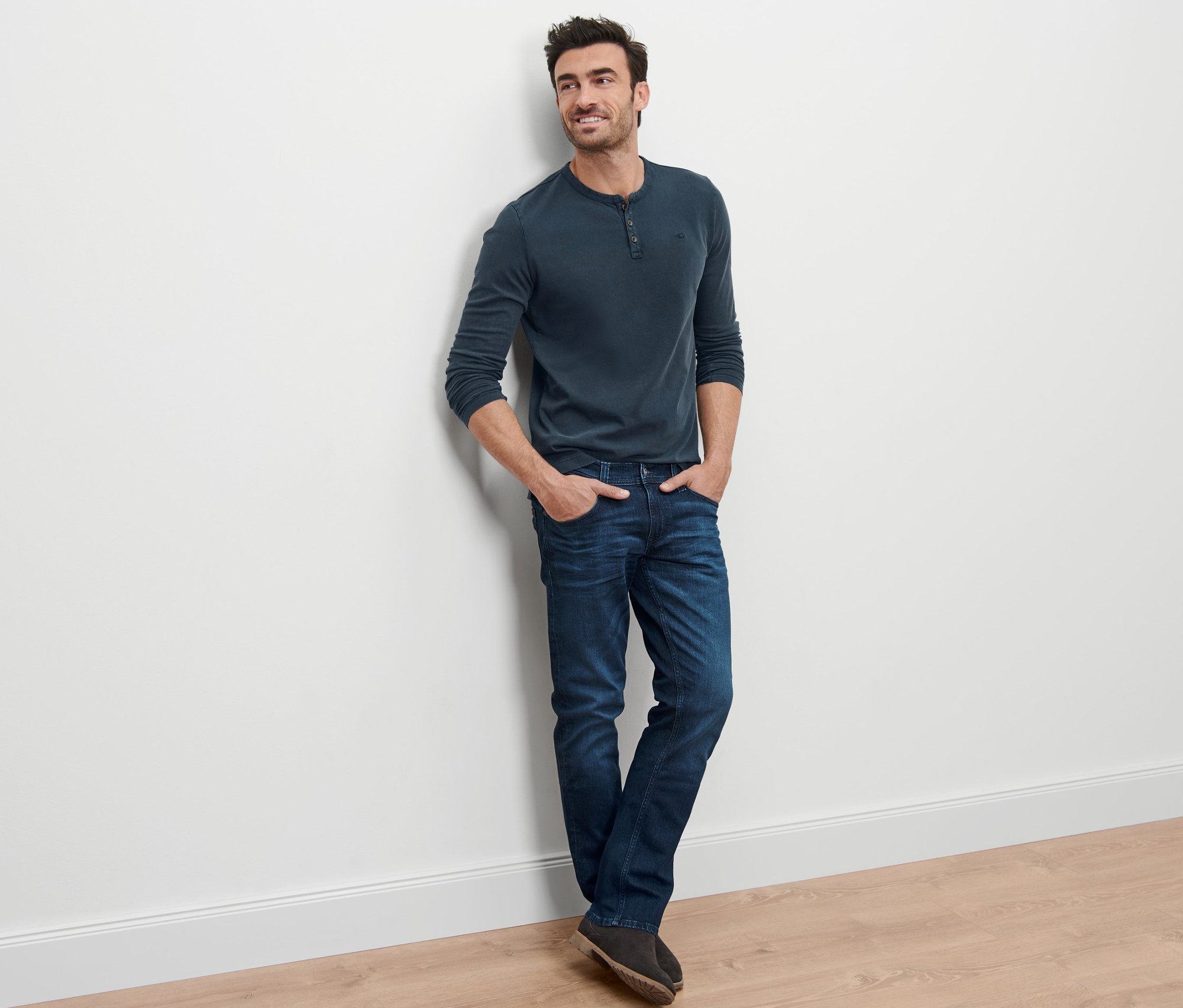Ein Mann lehnt an einer Wand und trägt Mustang Jeans »Oregon« und ein langärmliges Shirt.