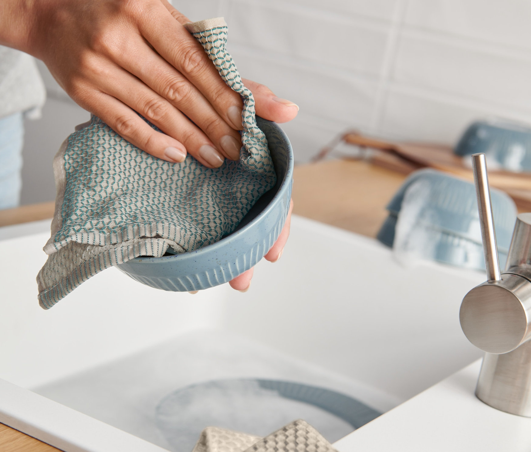 Eine Frau spült eine blaue Schüssel mit einem blauen Spültuch, auf der Arbeitsfläche liegen gefaltete 3 Spültücher.