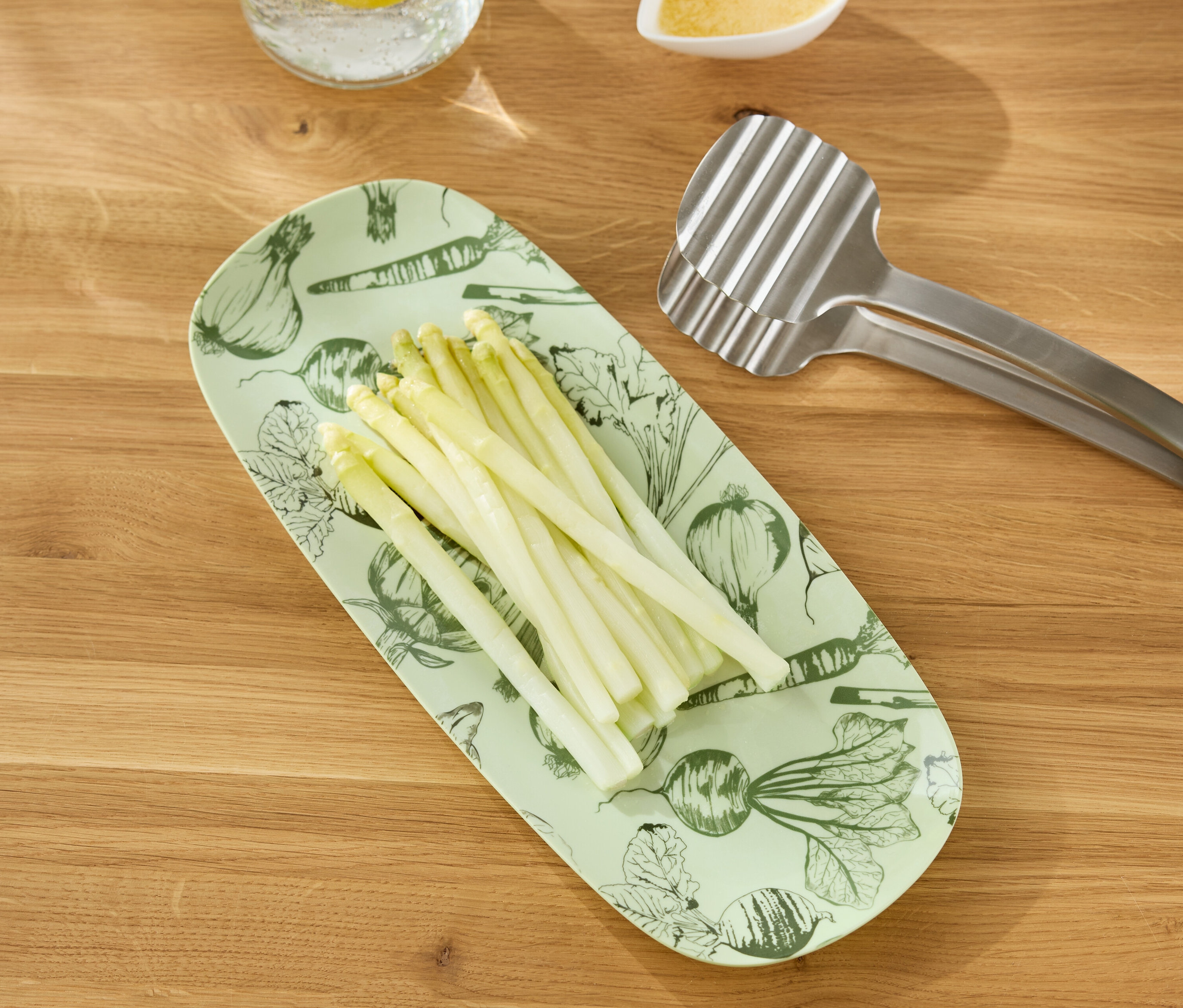 Weißer Spargel liegt auf einer länglichen, hellgrünen Servierplatte mit Gemüseaufdruck, daneben eine silberne Grillzange auf einem Holztisch.