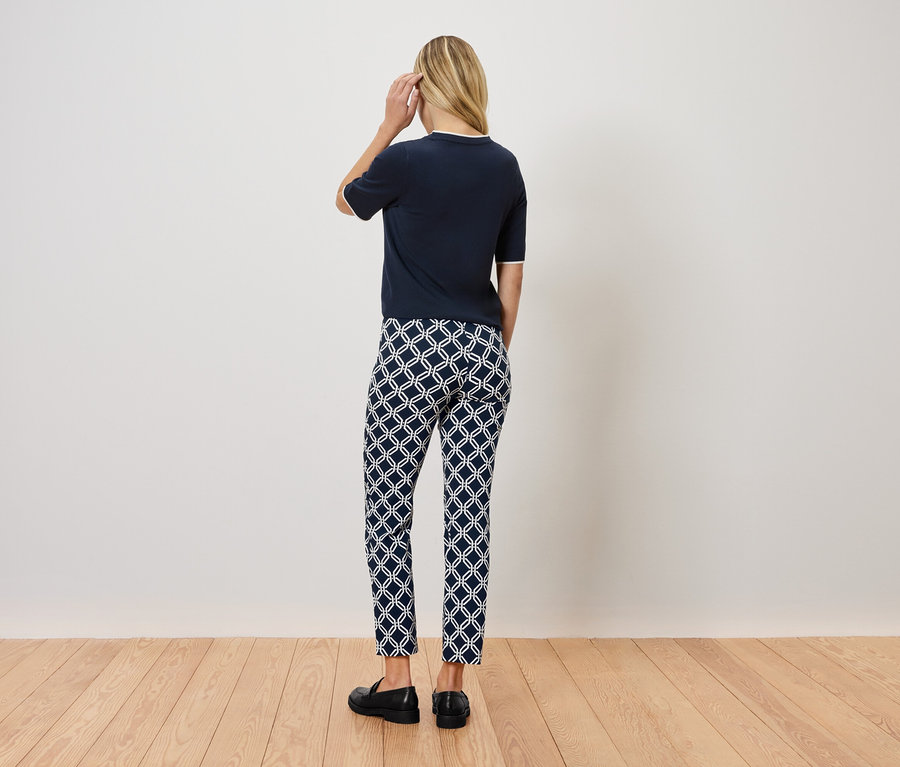 Frau steht mit dem Rücken zur Kamera und trägt ein dunkelblaues Oberteil, gemusterte Hosen und schwarze Schuhe.