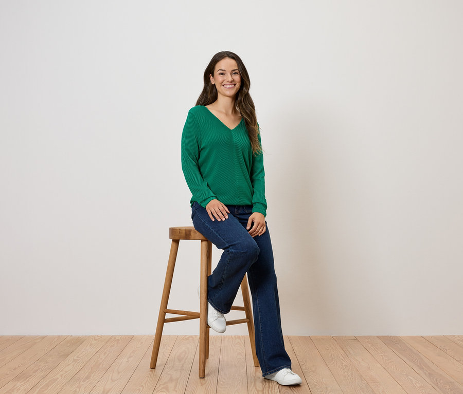 Eine Frau sitzt auf einem Holzhocker und trägt einen grünen Pullover und Jeans.