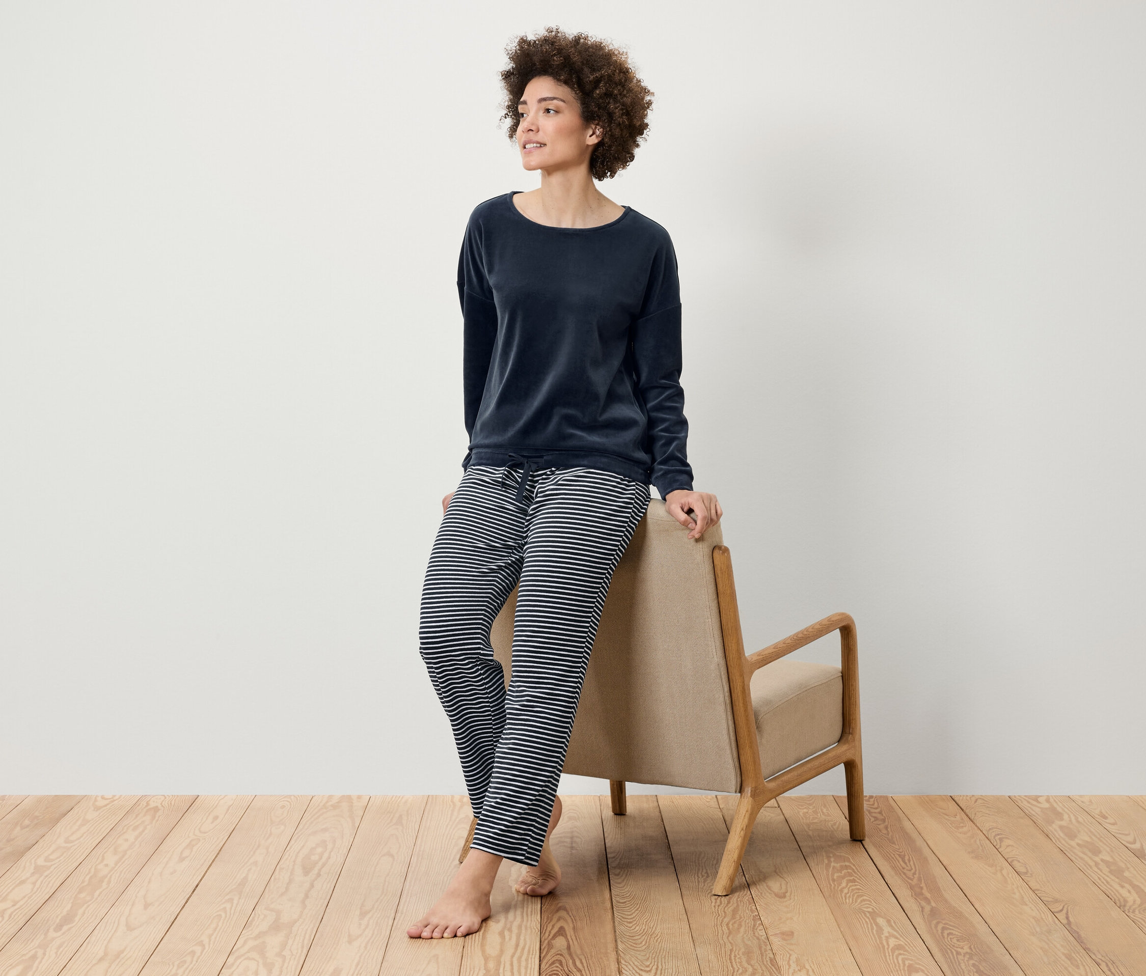 Frau mit Afro-Frisur posiert in einem marineblauen Samttop und einer gestreiften Hose, die sich an einen beigen Sessel auf einem Holzboden lehnt.