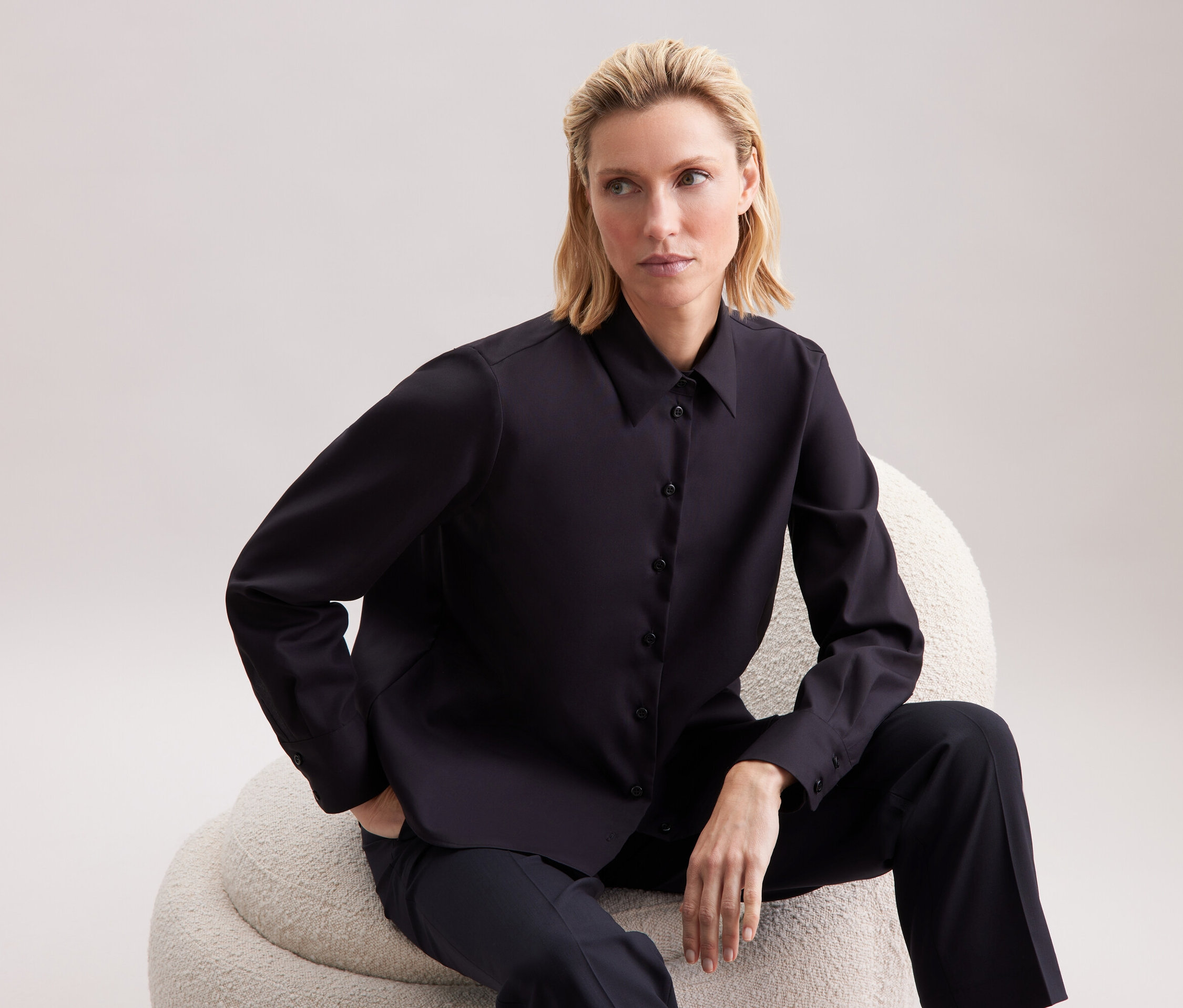 Blonde Frau sitzt auf einem beigen Sessel und trägt ein schwarzes Hemd und eine Hose.