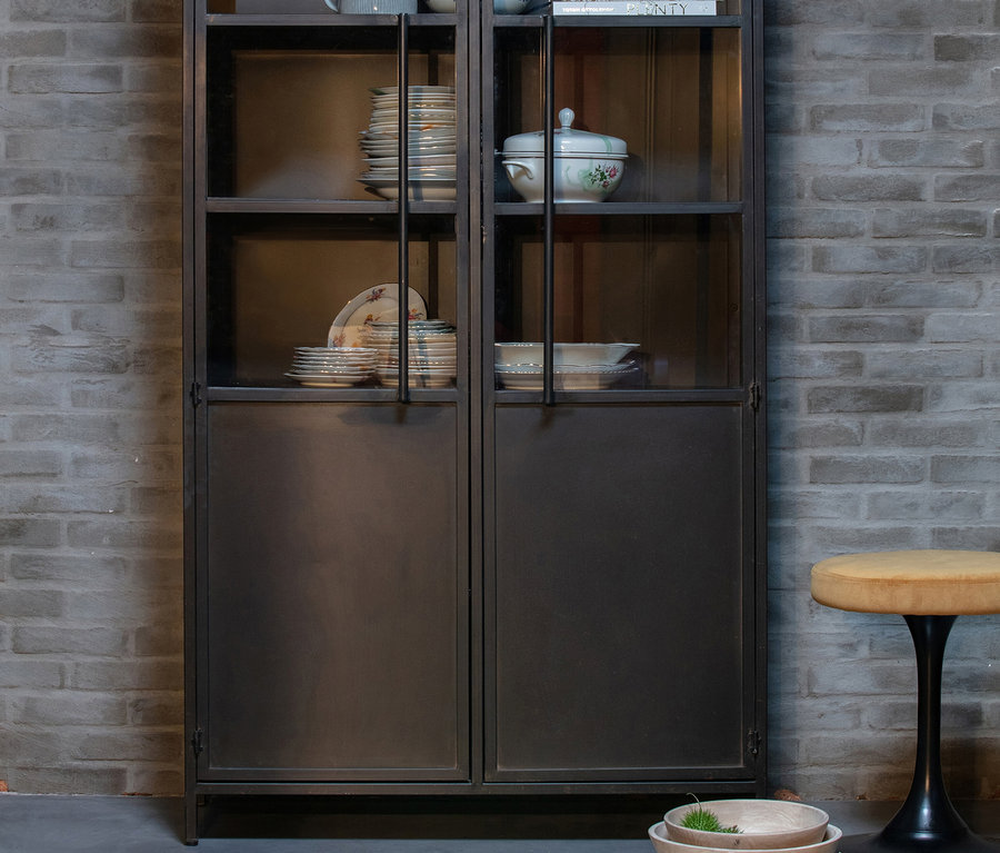 Khaki BePureHome Vitrine »Exhibit« aus Metall steht an einer Ziegelwand. In der Vitrine befinden sich Teller und Schüsseln.